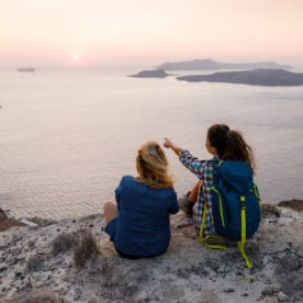 Back view of relaxed women resting on a break from hiking above the sea. One of them is aiming at distance. Copy space.