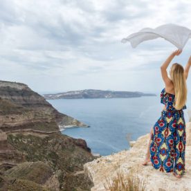 Horizontal color rear view image of young woman holding white scarf on the wind, standing at the edge of the cliff and feeling harmony with the sea.