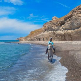A girl rides a grey horse along a volcanic beach on the Greek island of Santorini